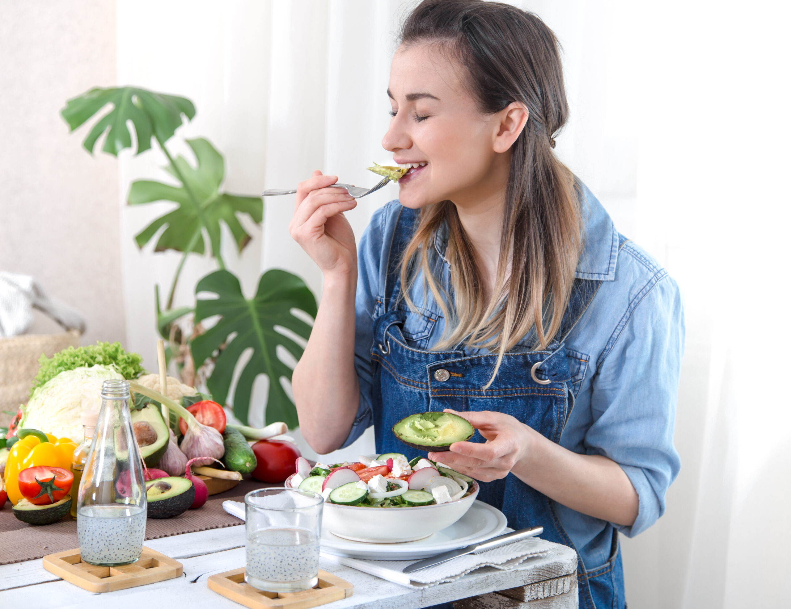 Young and happy woman eating salad with organic vegetables at the table on a light background , in denim clothes. The concept of a healthy home-made food.